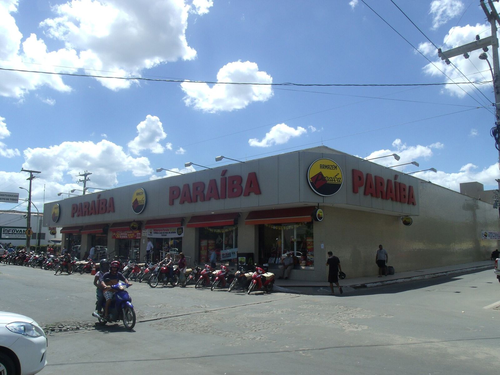 Esquina de rua com a loja do Armazém Paraíba em Piripiri (PI), fachada baixa com o letreiro vermelho "Paraíba" e o logotipo redondo amarelo, motos estacionadas na calçada e céu azul com nuvens