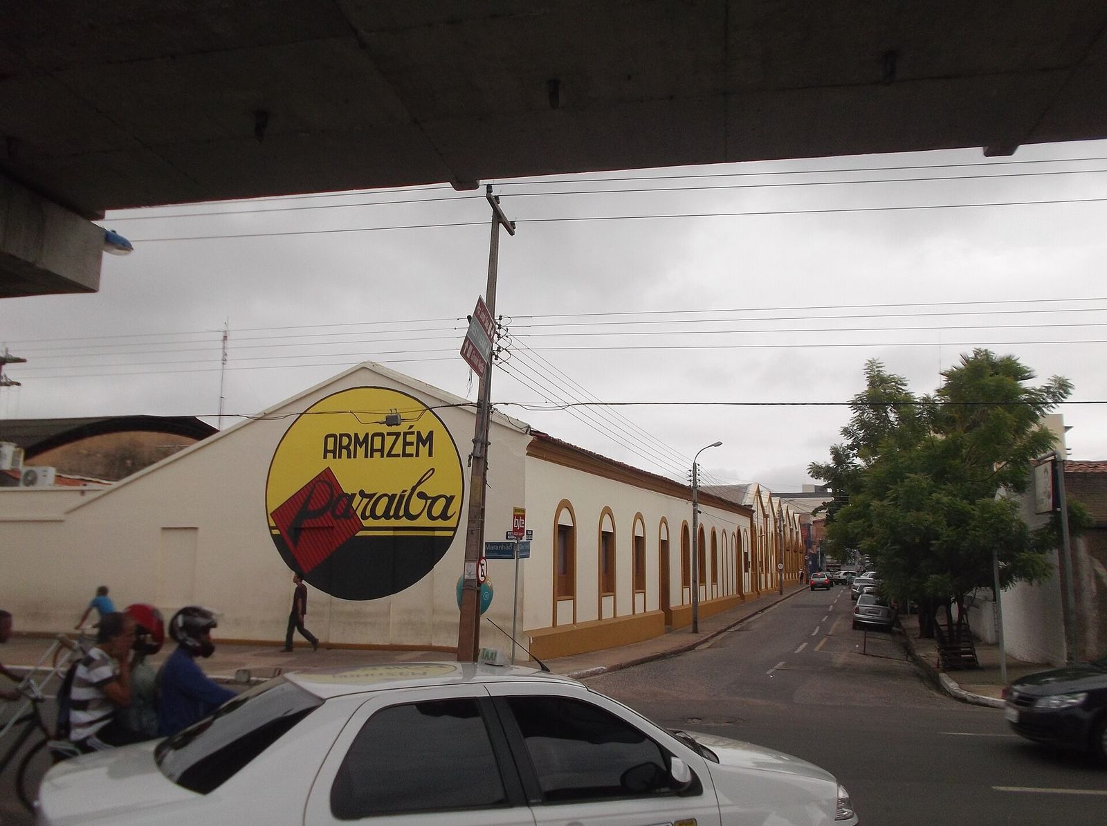 Prédio de paredes claras em esquina de Teresina, com grande letreiro redondo amarelo "Armazém Paraíba" na empena, rua em declive à direita e táxi branco em primeiro plano