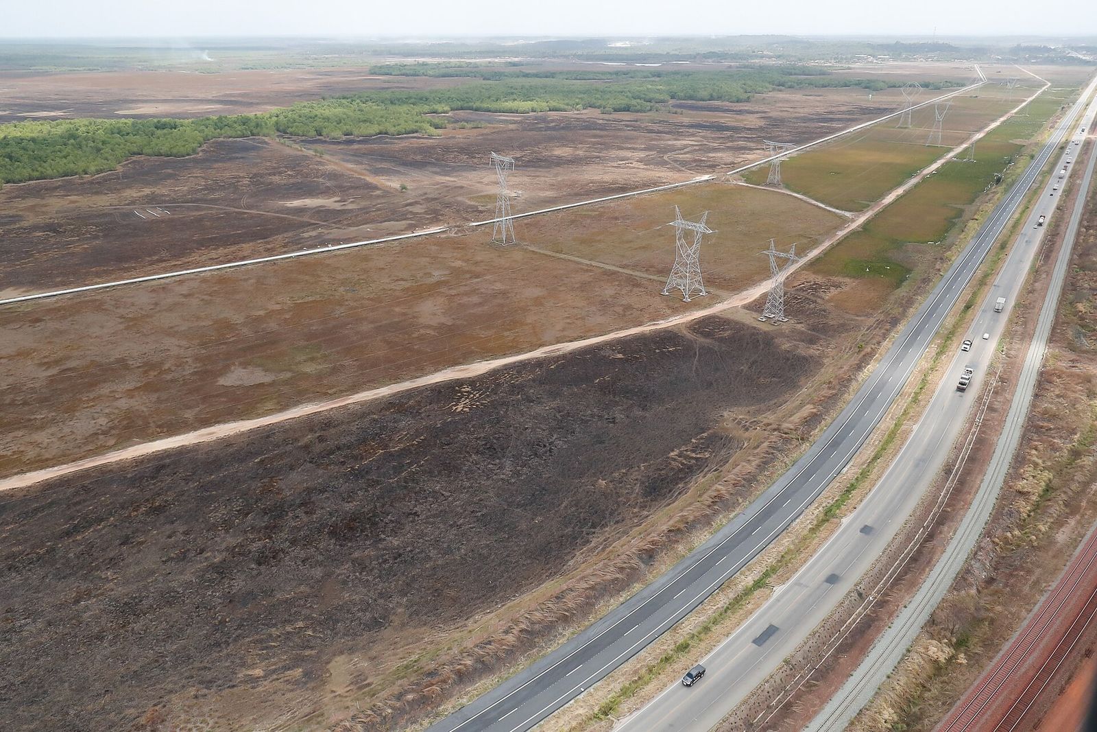 Vista aérea de uma rodovia de pista dupla ao lado de uma ferrovia, com linhas de transmissão e uma tubulação sobre uma planície alagável de capim e manguezal ao fundo