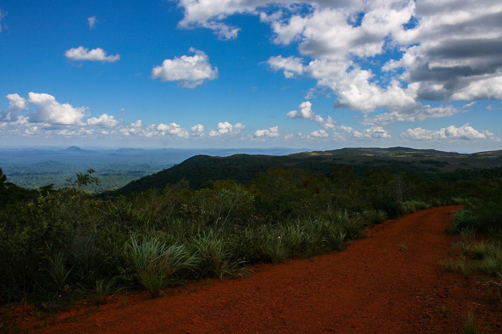 Estrada de terra vermelha entre vegetação baixa no alto da serra, com morros cobertos de floresta ao fundo e céu azul com nuvens