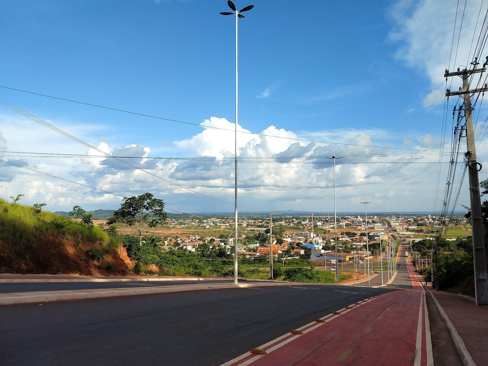 Avenida asfaltada com ciclovia vermelha descendo em direção à cidade, com casas e árvores ao fundo