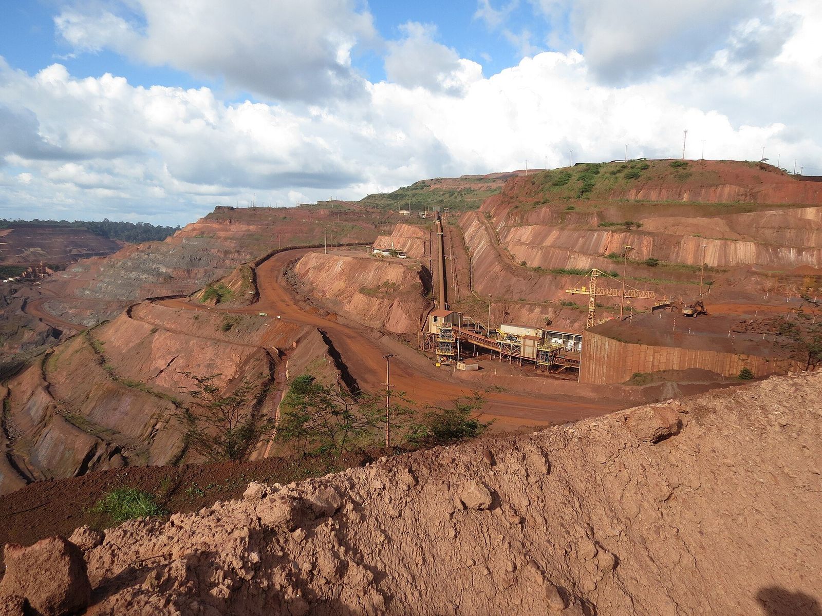 Cava de mina a céu aberto com bancadas de terra avermelhada e uma instalação industrial com correias transportadoras