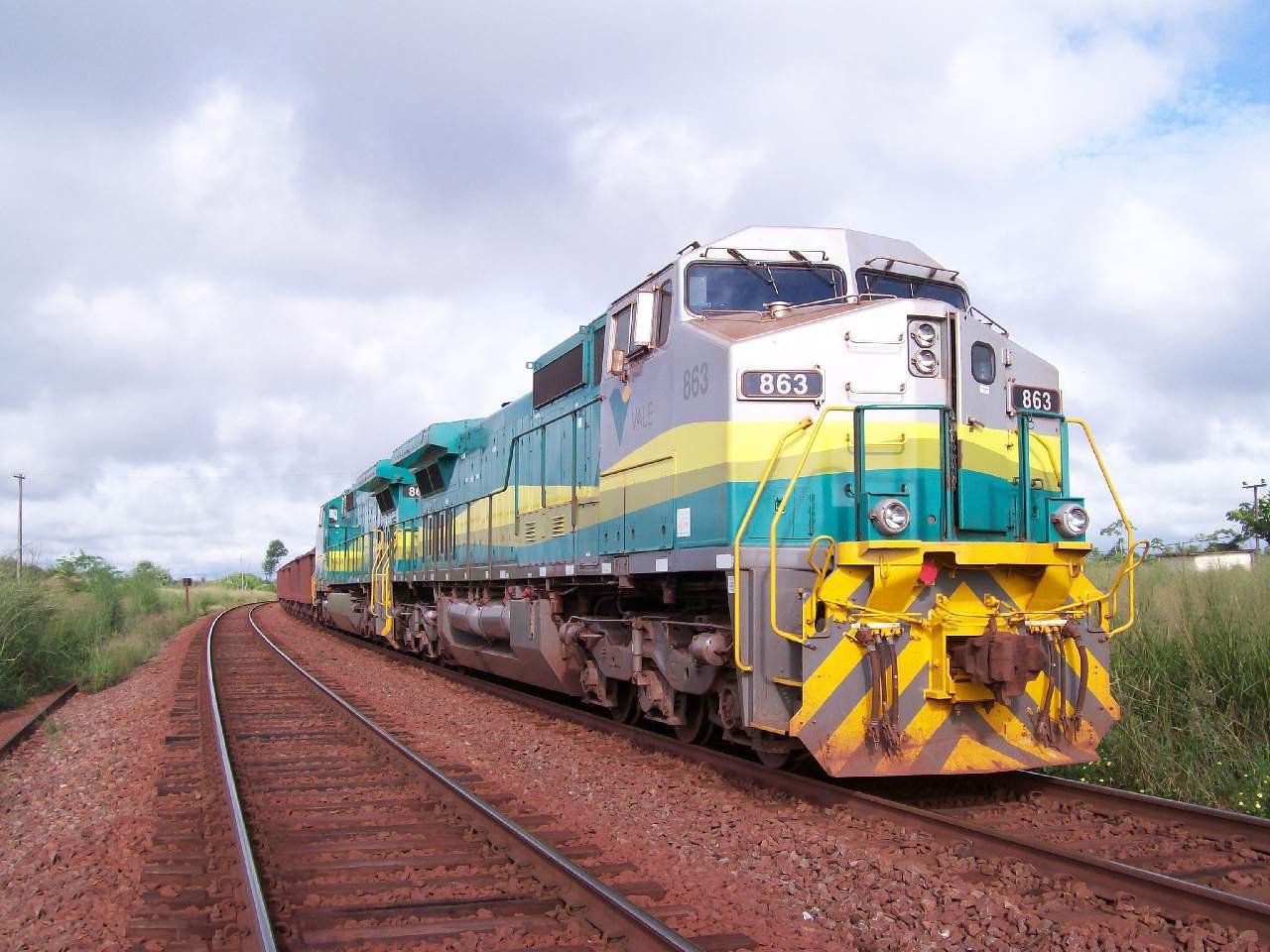Locomotiva verde, amarela e cinza sobre trilhos com lastro avermelhado, entre capim alto