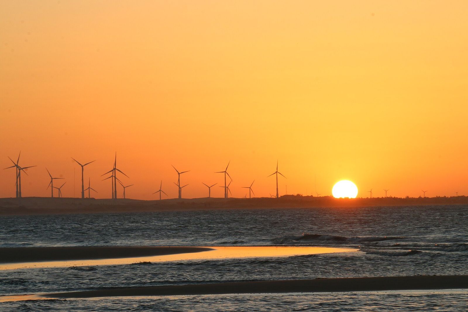Pôr do sol alaranjado sobre a praia do Pecém, com uma fileira de turbinas eólicas no horizonte e a maré baixa refletindo a luz