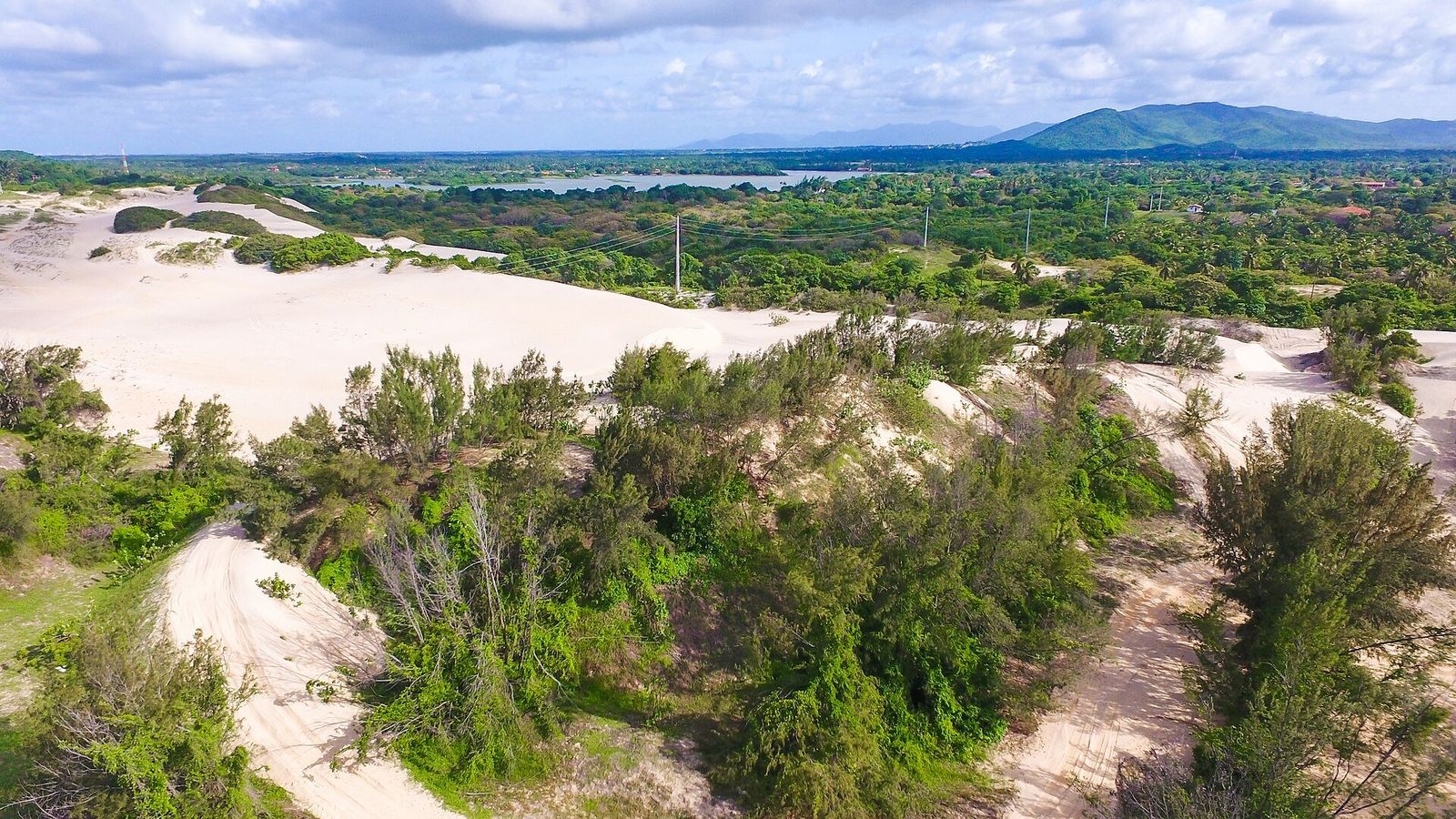 Vista aérea de dunas brancas cobertas em parte por vegetação, com uma lagoa e serras ao fundo