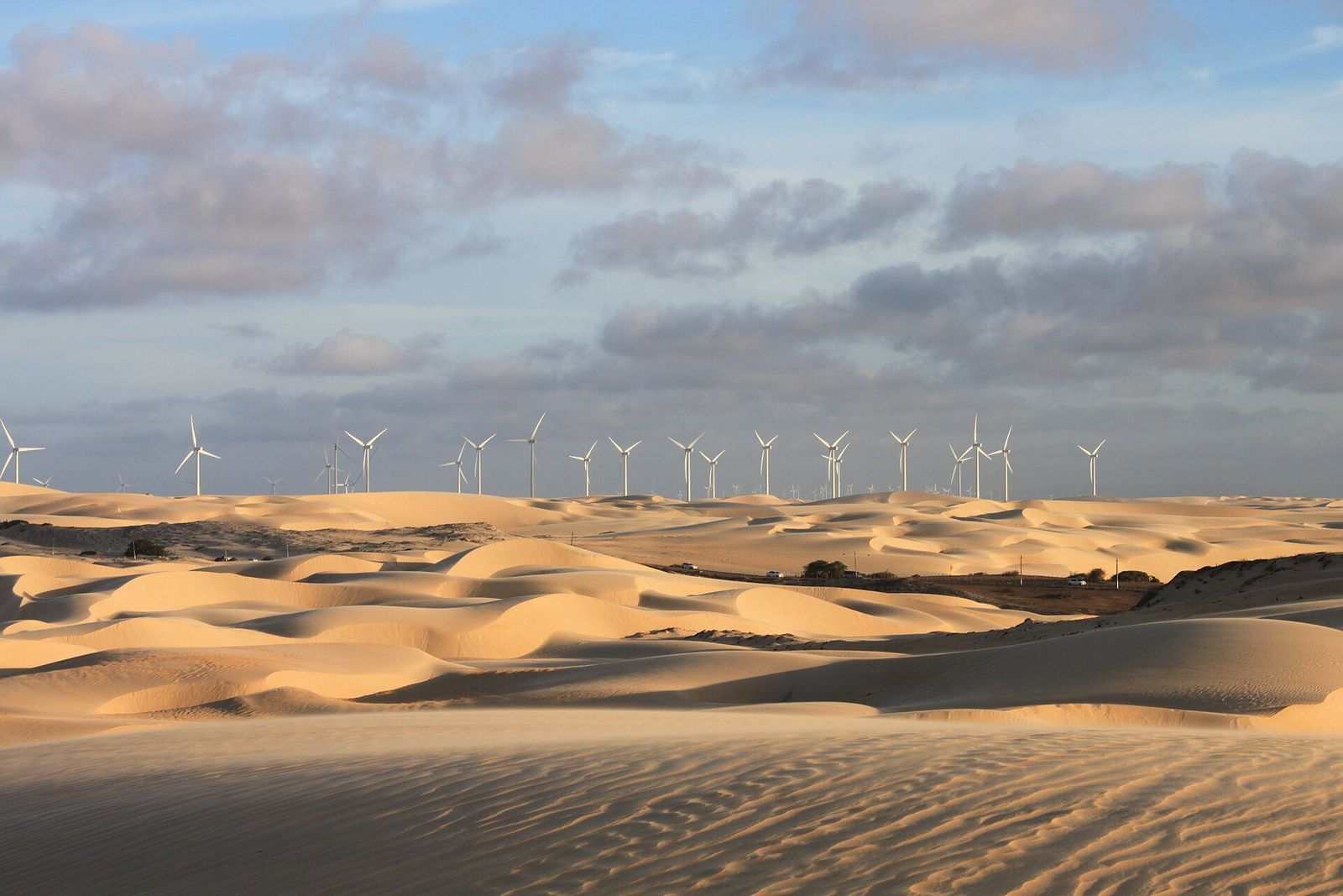 Fileira de turbinas eólicas no horizonte, atrás de dunas de areia sob céu com nuvens