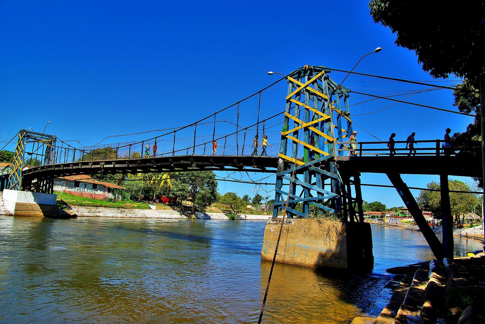 Ponte pênsil de madeira pintada de azul e amarelo sobre o rio, com pessoas atravessando, margens com casas e árvores ao fundo e céu azul