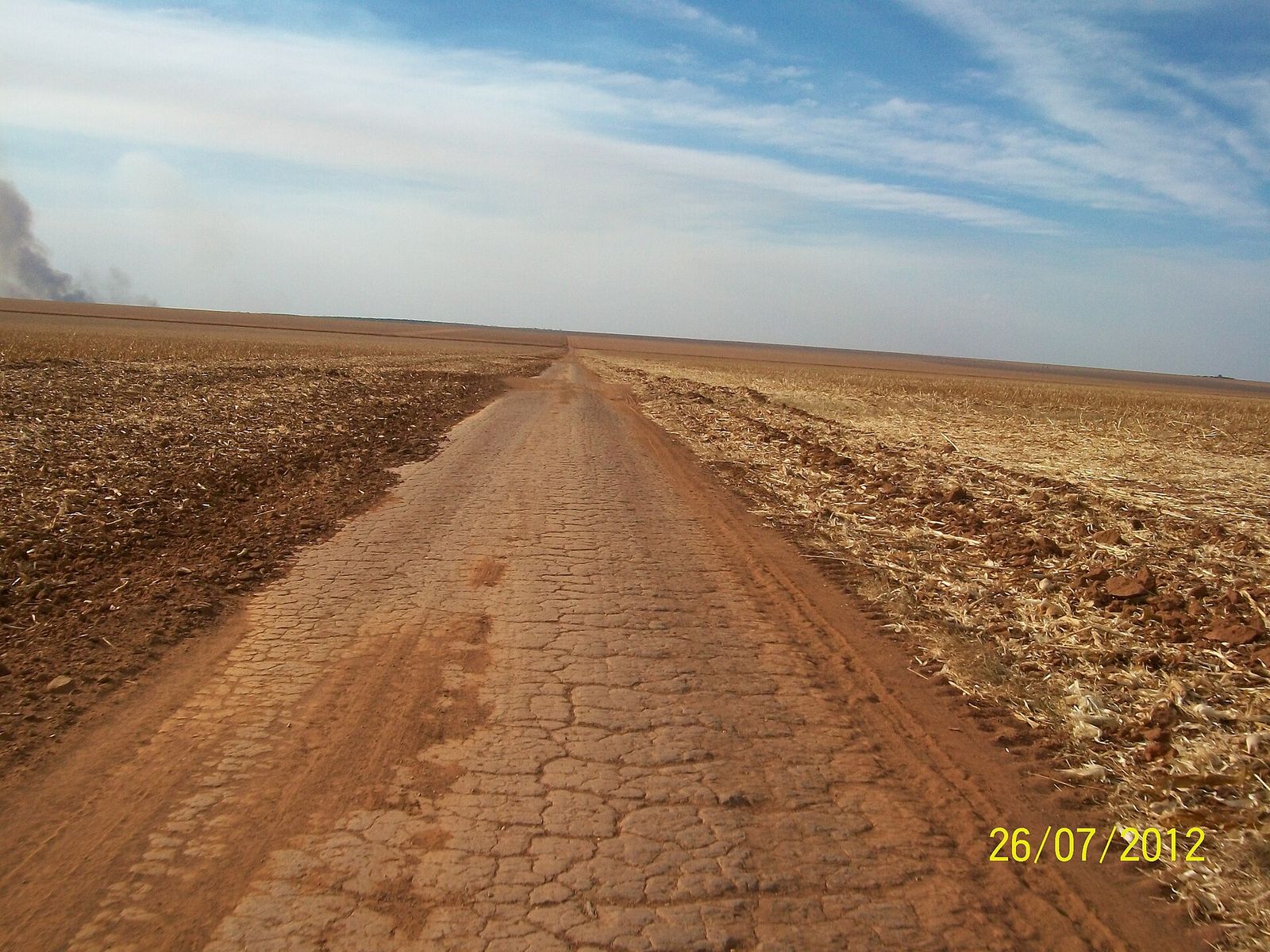 Estrada de terra vermelha entre campos de lavoura em terreno plano, sob céu azul com nuvens finas