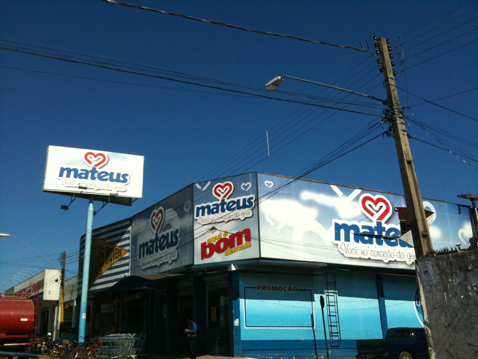Esquina com fachada azul e branca de uma loja Mateus, placa alta com o coração vermelho da marca, postes e fios