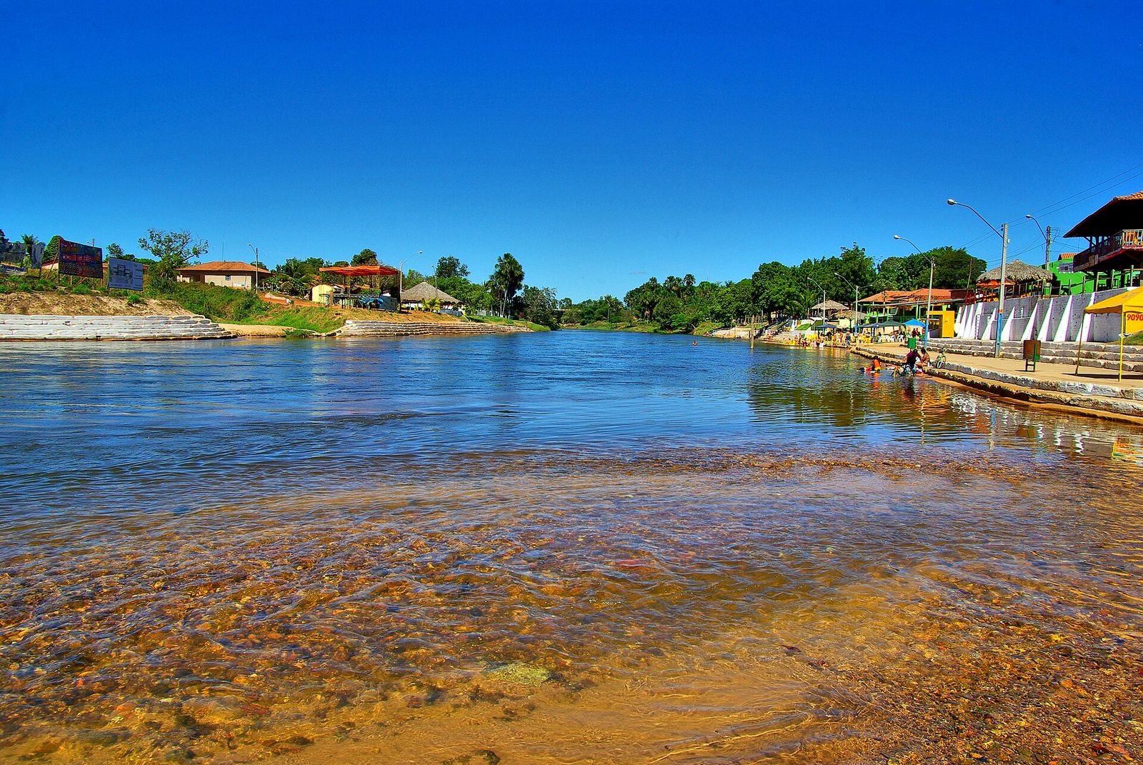 Rio de águas claras e margens com casas, árvores e quiosques sob céu azul