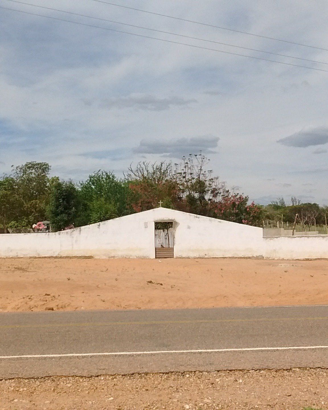Beira de rodovia asfaltada com terreno de terra batida e, ao fundo, o muro branco de um cemitério com árvores atrás, sob céu nublado
