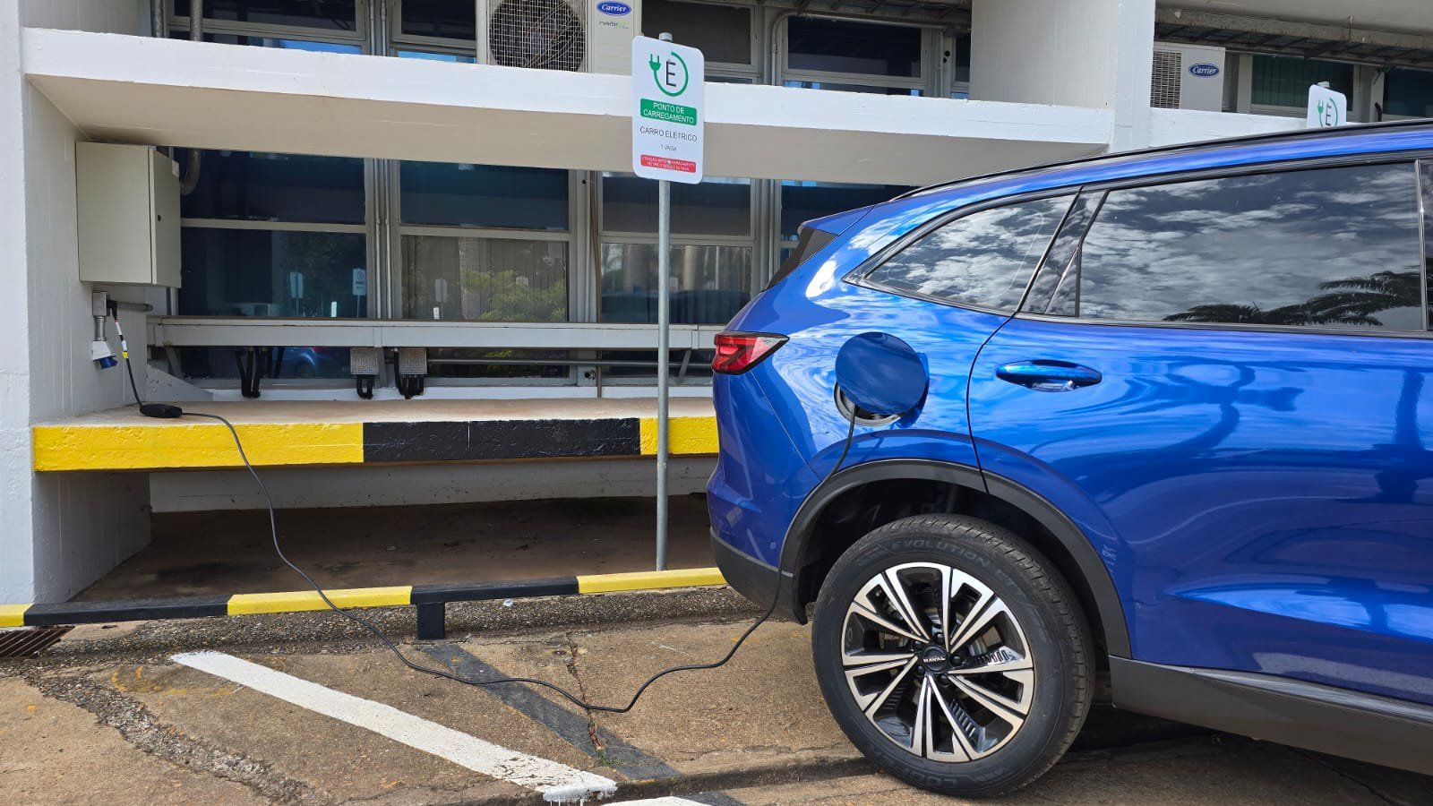 Carro elétrico azul estacionado ligado por cabo a um ponto de recarga diante de um prédio