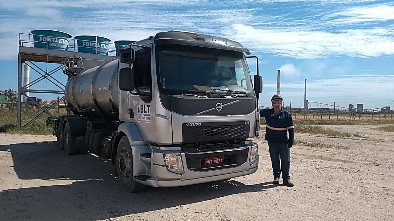 Caminhão-pipa prateado estacionado em terreno de terra batida, com um trabalhador de uniforme ao lado