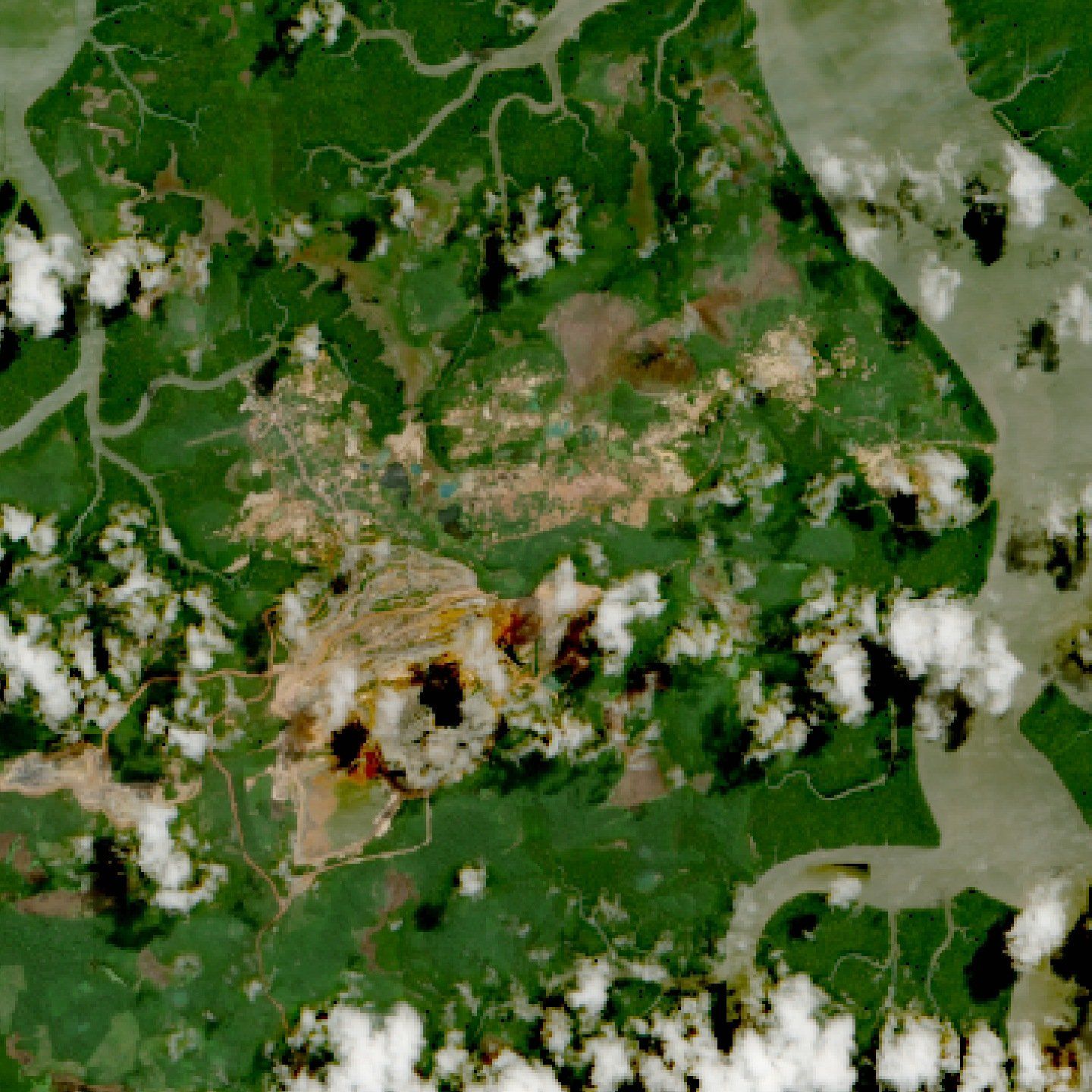 Imagem de satélite com a área da mina Aurizona: cavas e solo exposto em tons de bege e marrom cercados por mata e manguezais, com nuvens esparsas