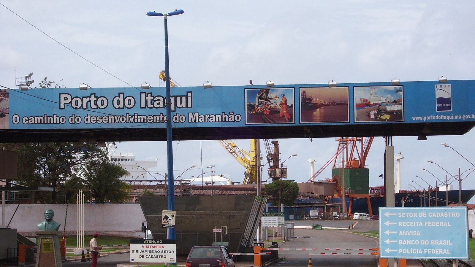 Pórtico azul com a inscrição Porto do Itaqui, o caminho do desenvolvimento do Maranhão, sobre a entrada do porto, com guindastes e um navio ao fundo