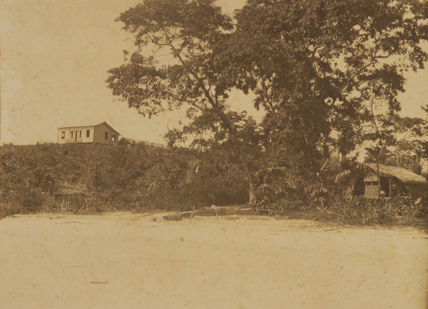 Praia de areia com mata, uma casa no morro e uma palhoça, em foto sépia