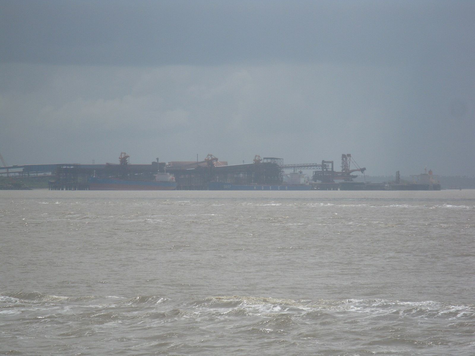 Estruturas de carregamento de navios na margem da baía, vistas de longe, sob céu carregado