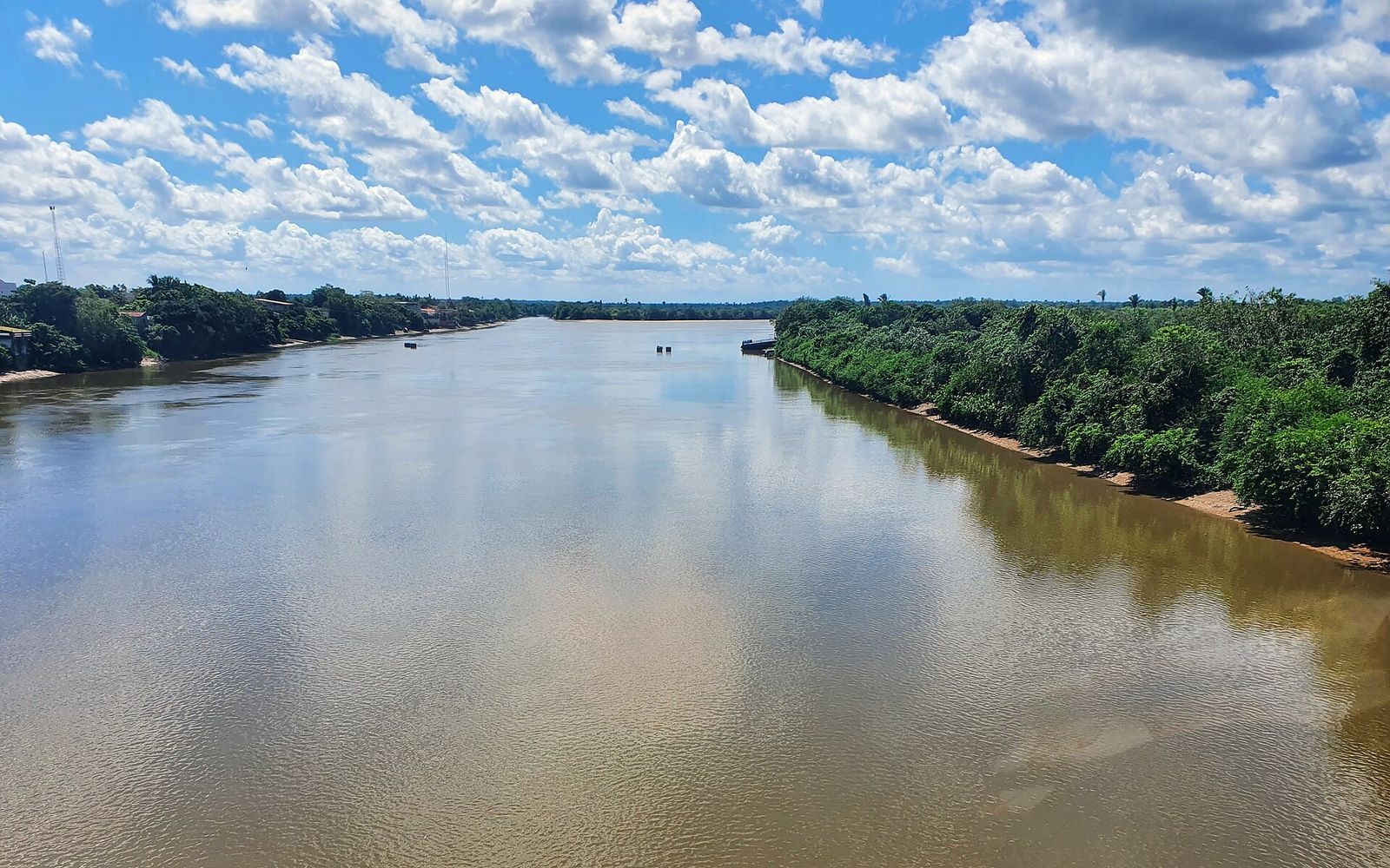Rio largo e de águas barrentas entre margens de mata, sob céu azul com nuvens
