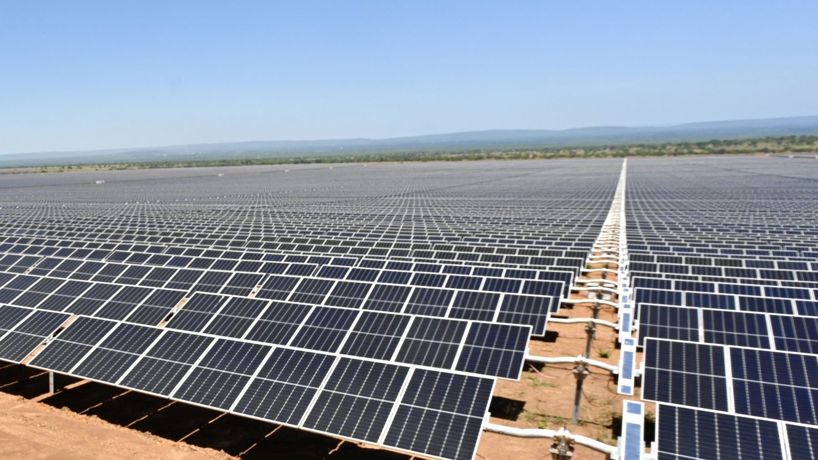 Fileiras de painéis solares azuis sobre chão de terra avermelhada, estendendo-se até o horizonte sob céu azul