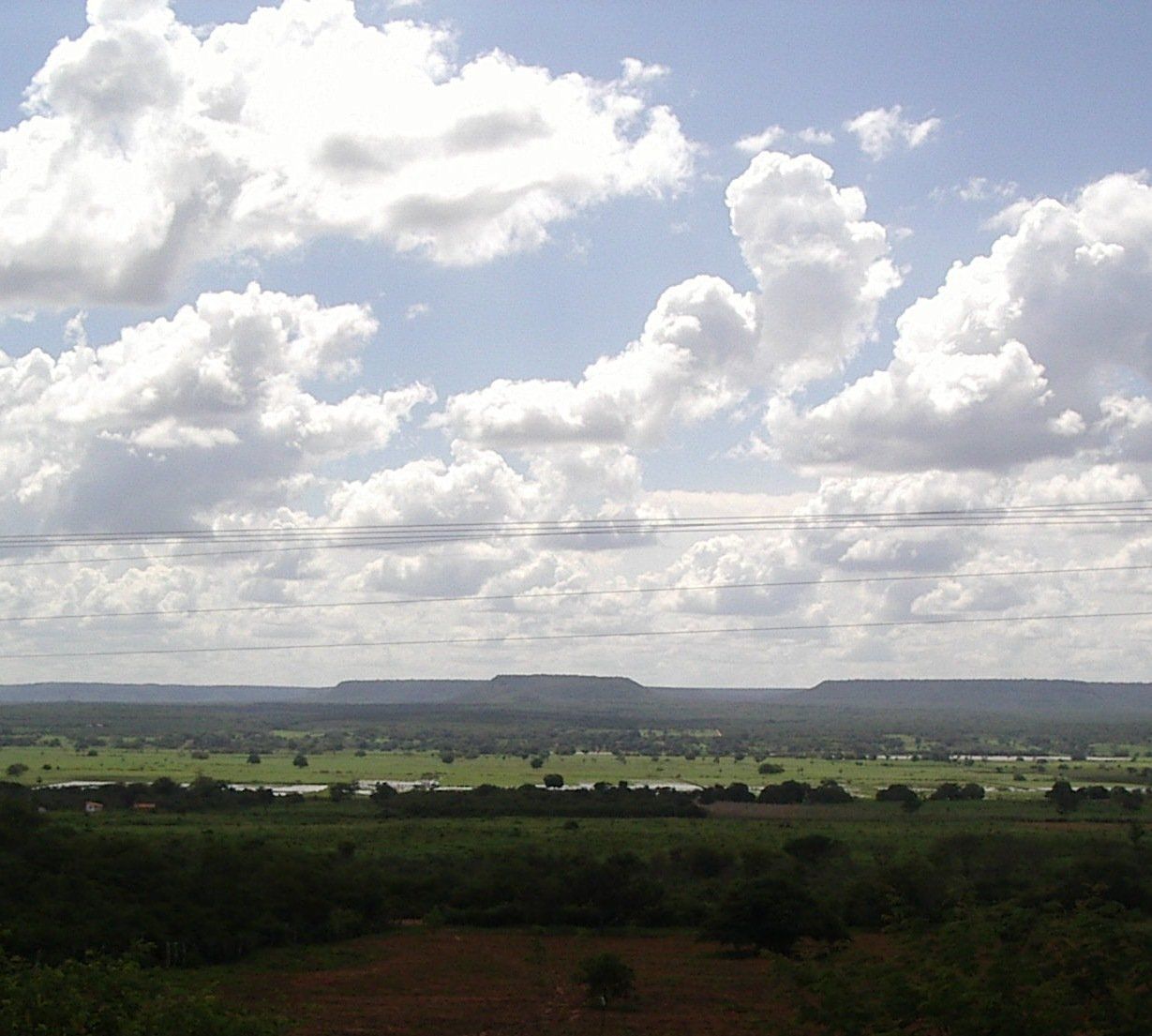 Vista do alto de um vale plano com vegetação verde de cerrado, chapadas no horizonte e céu com muitas nuvens brancas