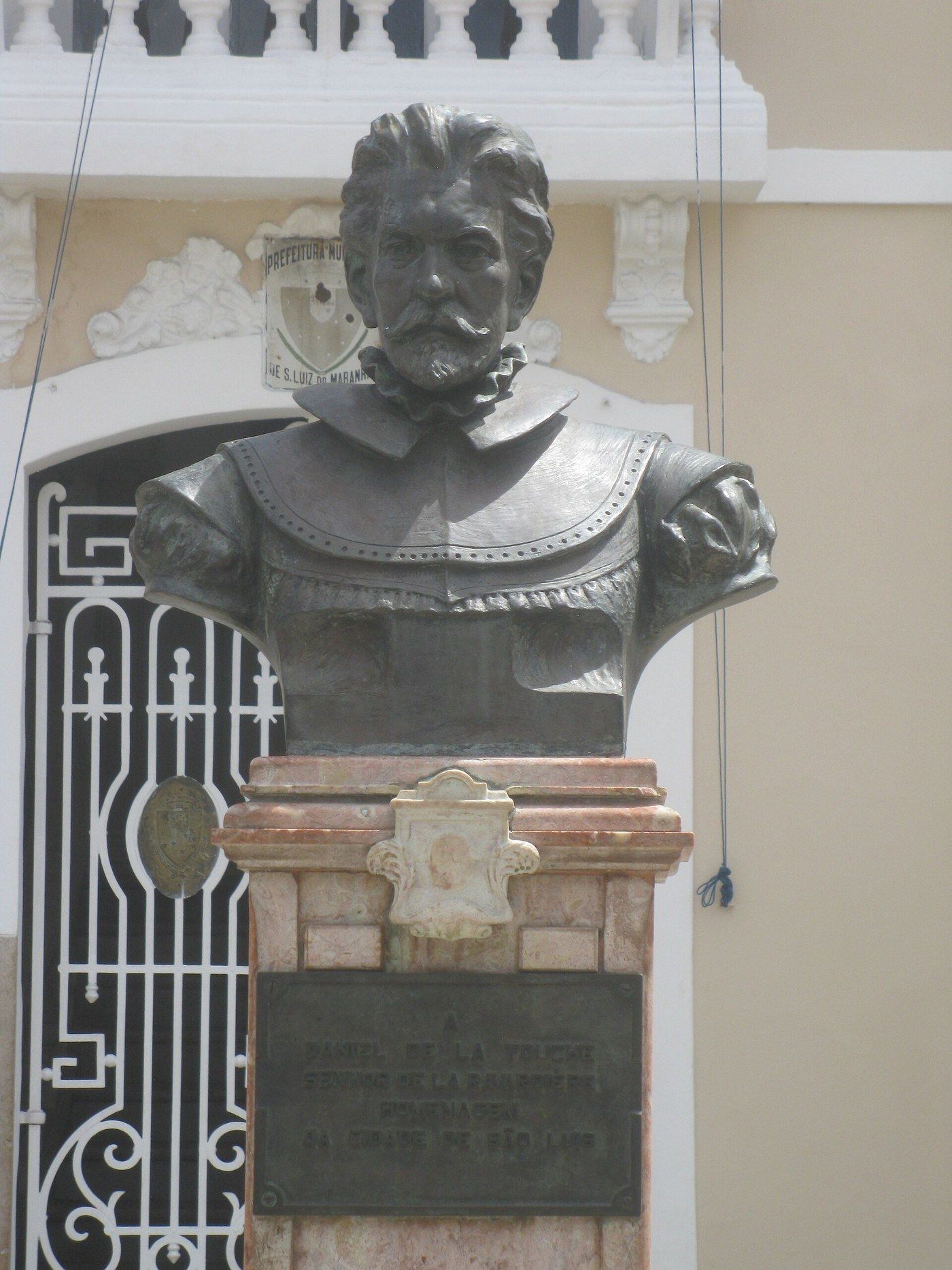 Busto de bronze de um homem de bigode e barba, com gola de renda, sobre pedestal de mármore rosado com placa de homenagem