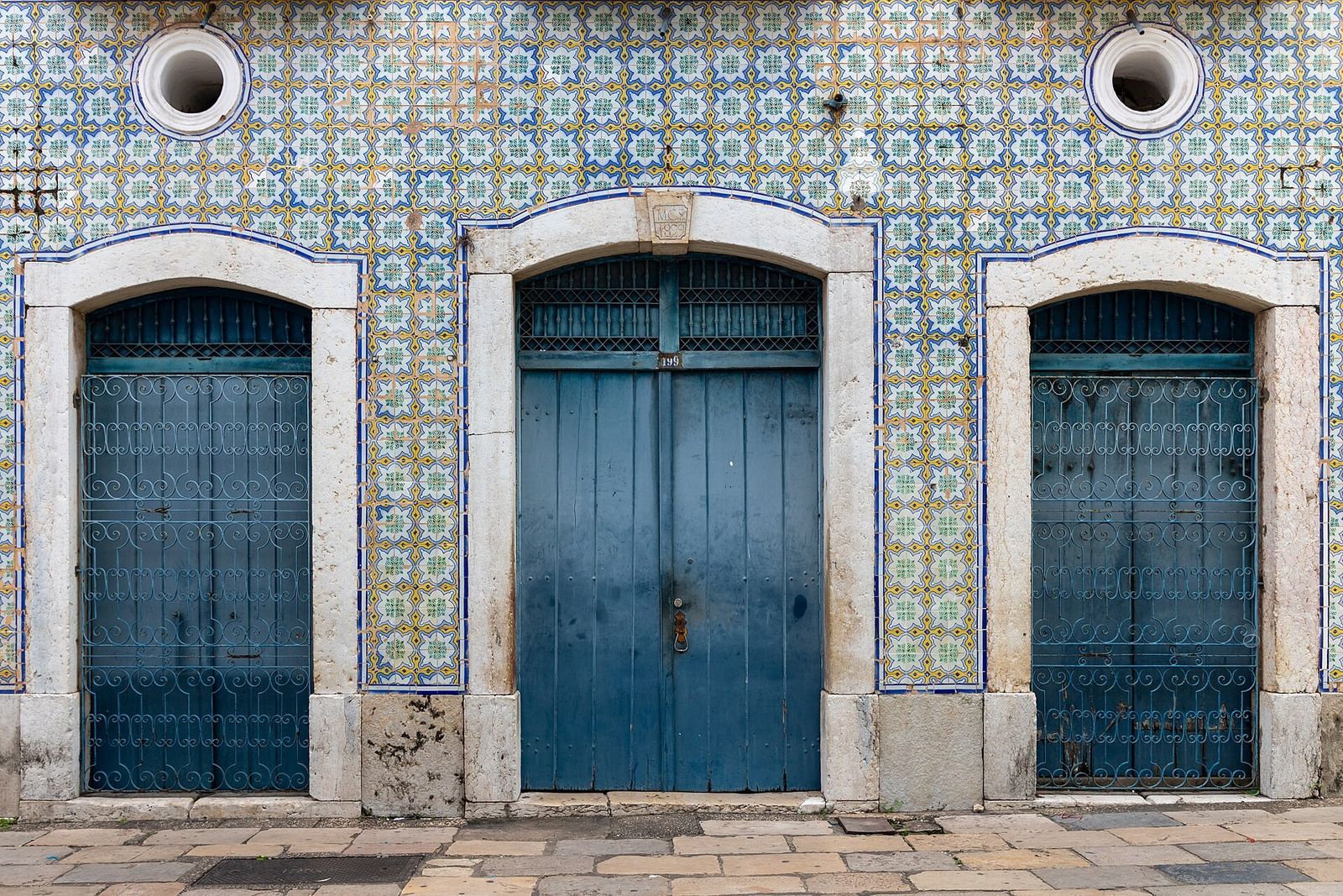 Fachada de sobrado coberta de azulejos azuis, amarelos e verdes, com três portas de madeira azul e grades de ferro entre molduras de pedra clara
