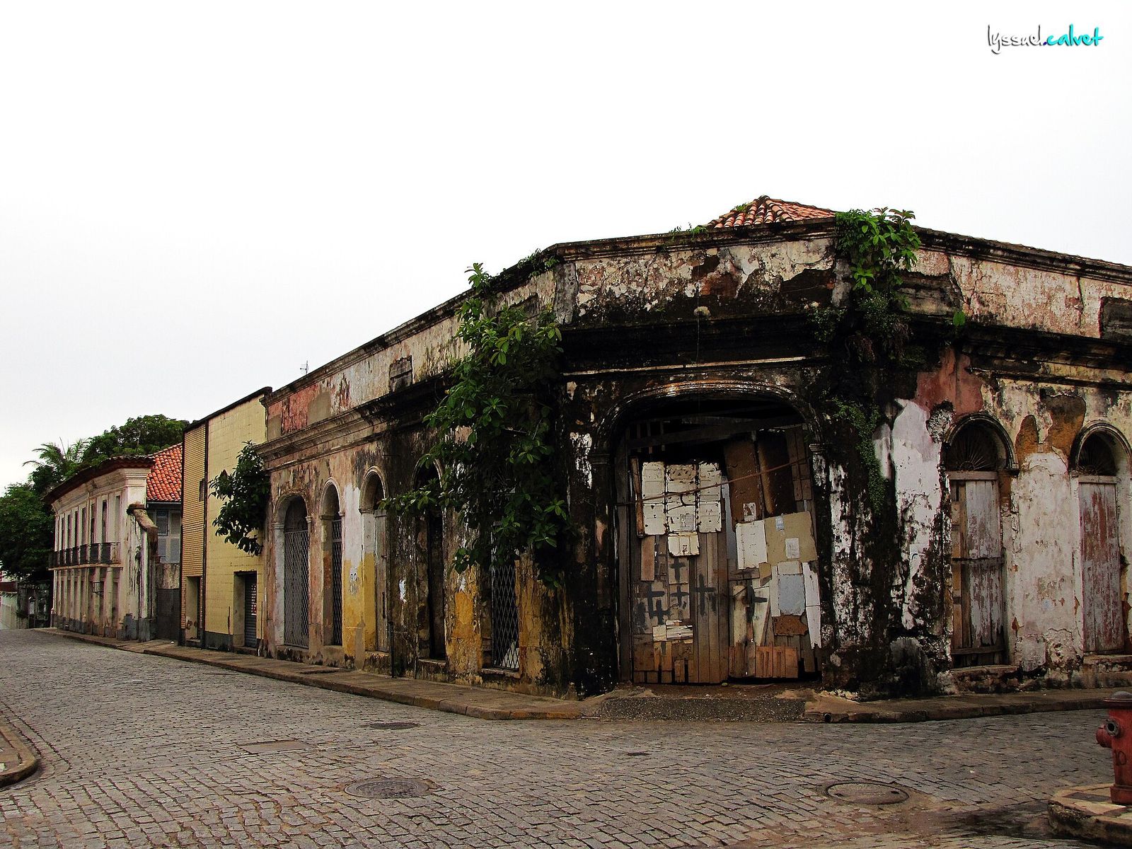Casarão de esquina com fachada manchada, mato nas cornijas e portas fechadas com tábuas, numa rua de paralelepípedos