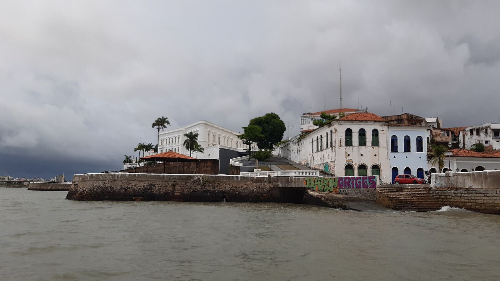 Palácio branco de vários andares sobre um muro de pedra à beira da baía, sob céu carregado, com casarões coloniais à direita