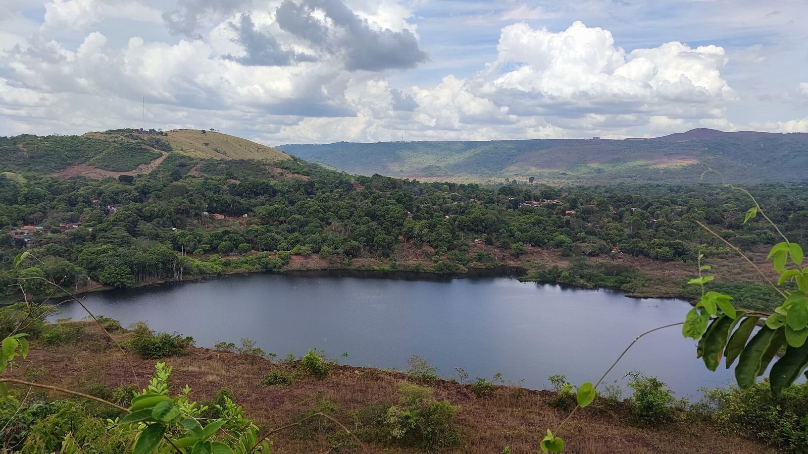 Lago escuro ocupando a cava do antigo garimpo de Serra Pelada, cercado por morros cobertos de mata, com casas da vila na margem oposta e céu de nuvens carregadas
