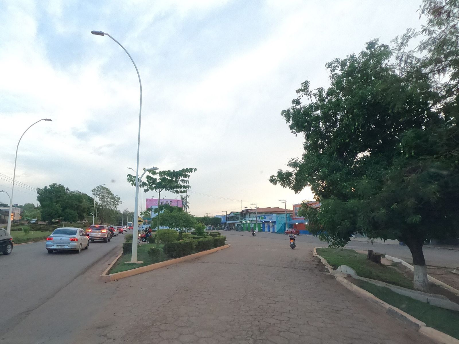 Avenida asfaltada em Curionópolis com canteiro central, postes de iluminação, carros e comércio ao fundo, ao entardecer