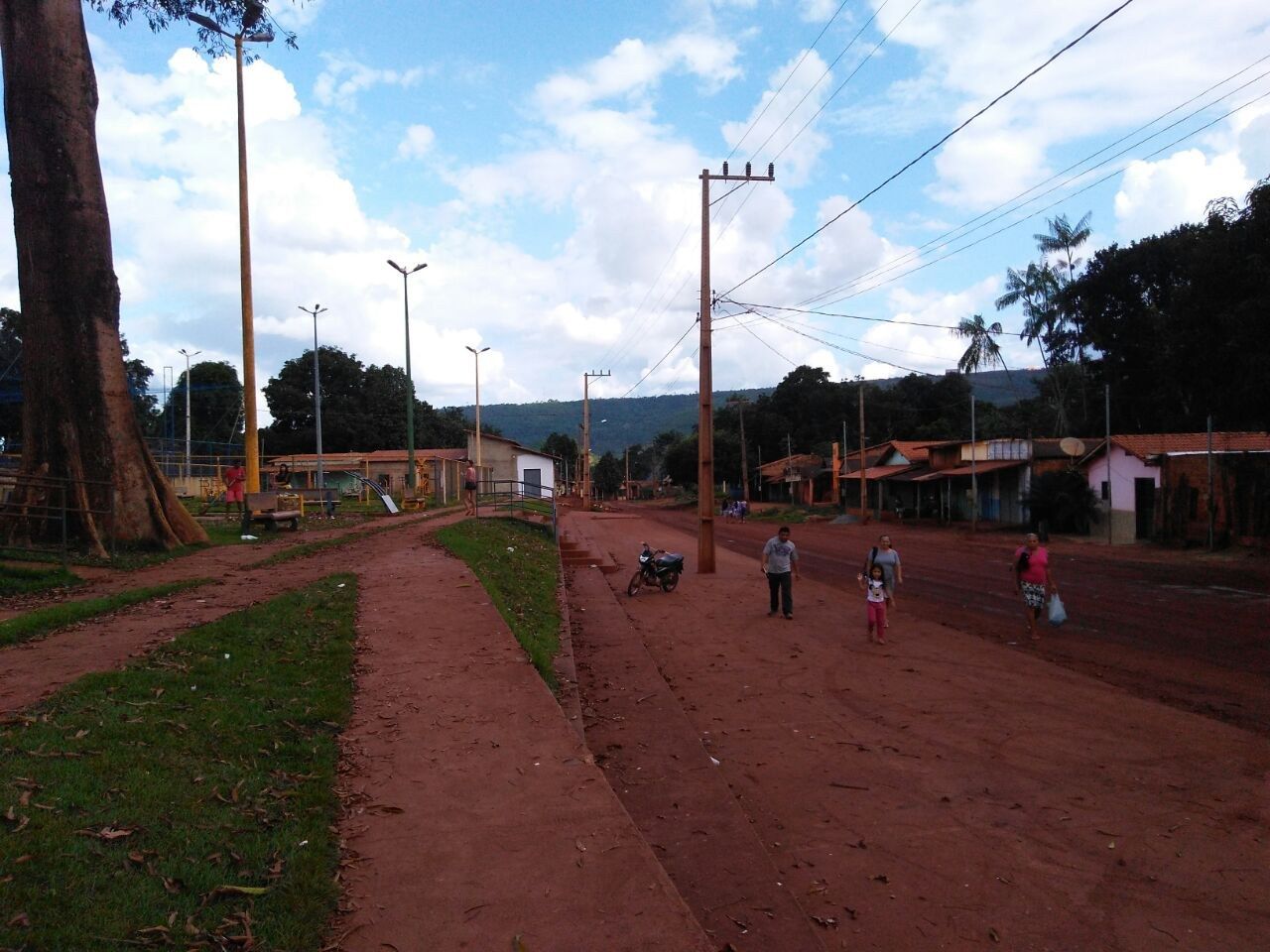 Rua de terra vermelha na vila de Serra Pelada, com praça à esquerda, postes, casas baixas e morro ao fundo