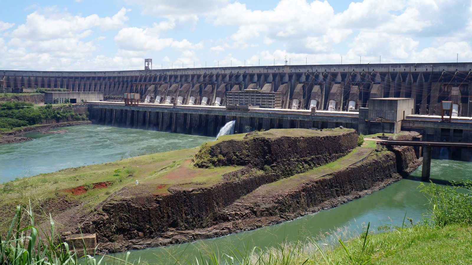 Vista da barragem de Itaipu com o paredão de concreto ao fundo, as saídas das turbinas e rochas escuras em primeiro plano sob céu com nuvens
