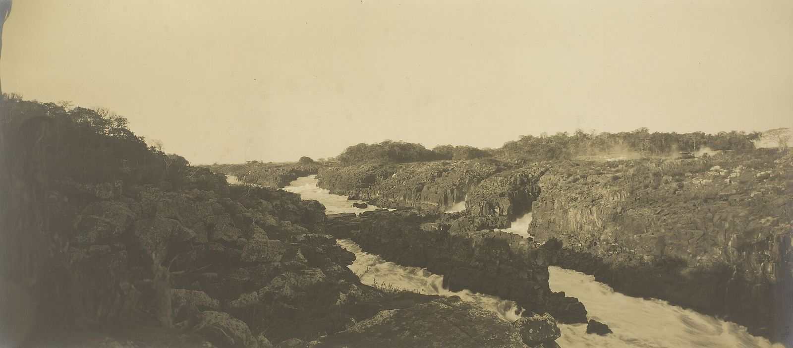 Fotografia antiga em sépia do rio Paraná estreitado entre blocos de rocha escura, com várias corredeiras e pequenas quedas brancas ao longo do canal