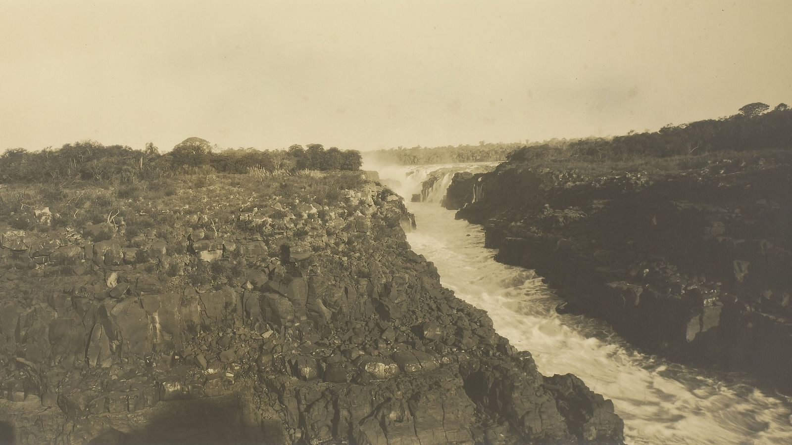 Fotografia antiga em tons de sépia do rio Paraná espremido entre paredões de rocha escura, com corredeiras brancas em primeiro plano e uma queda de água ao fundo, sob céu claro