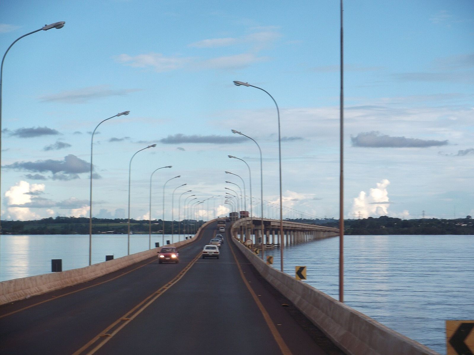 Ponte comprida sobre o rio Paraná, com postes de iluminação, carros na pista e águas calmas dos dois lados sob céu azul com nuvens