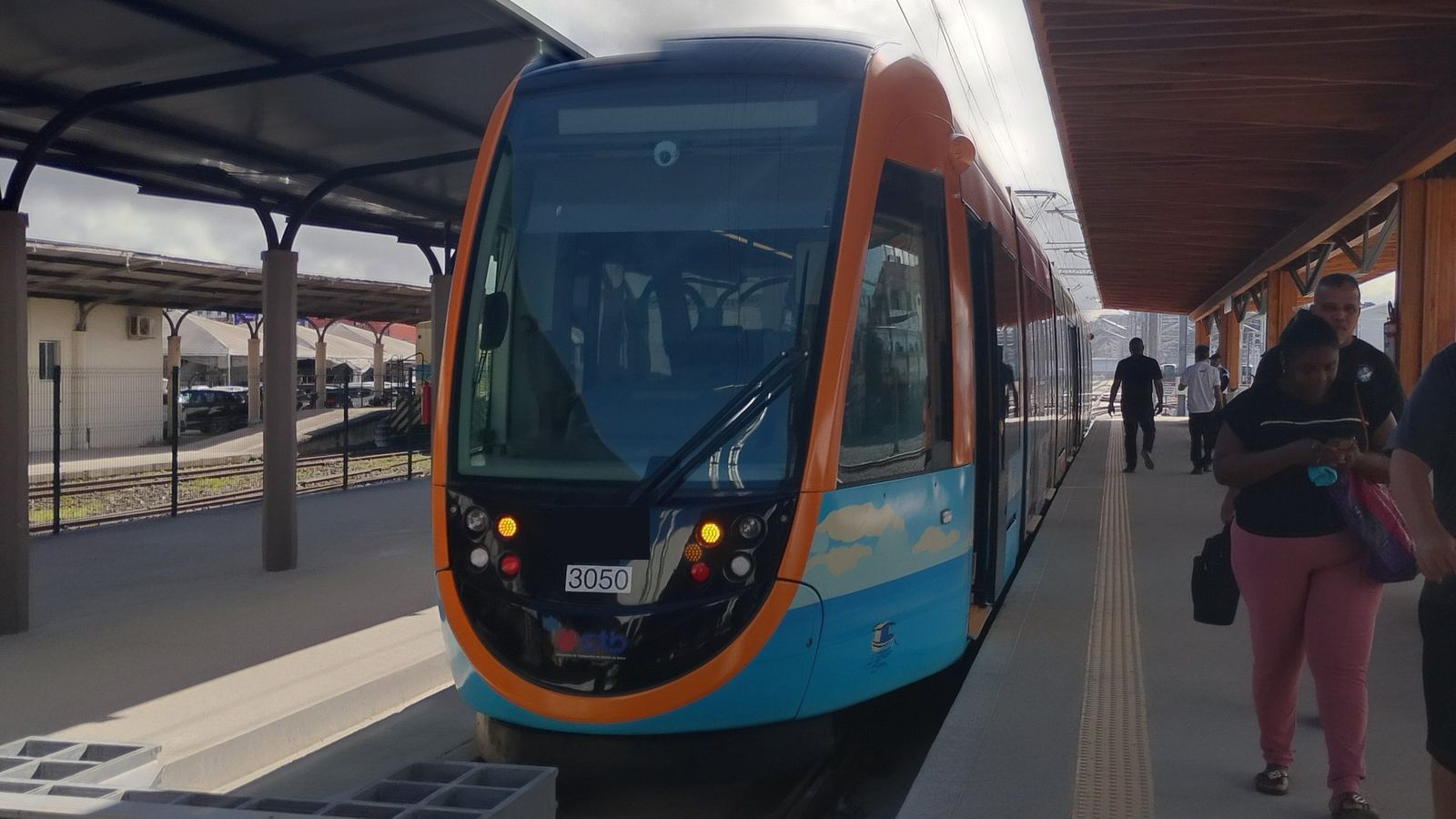 Trem do VLT, azul e laranja, parado na plataforma coberta da Estação Calçada, com passageiros caminhando ao lado