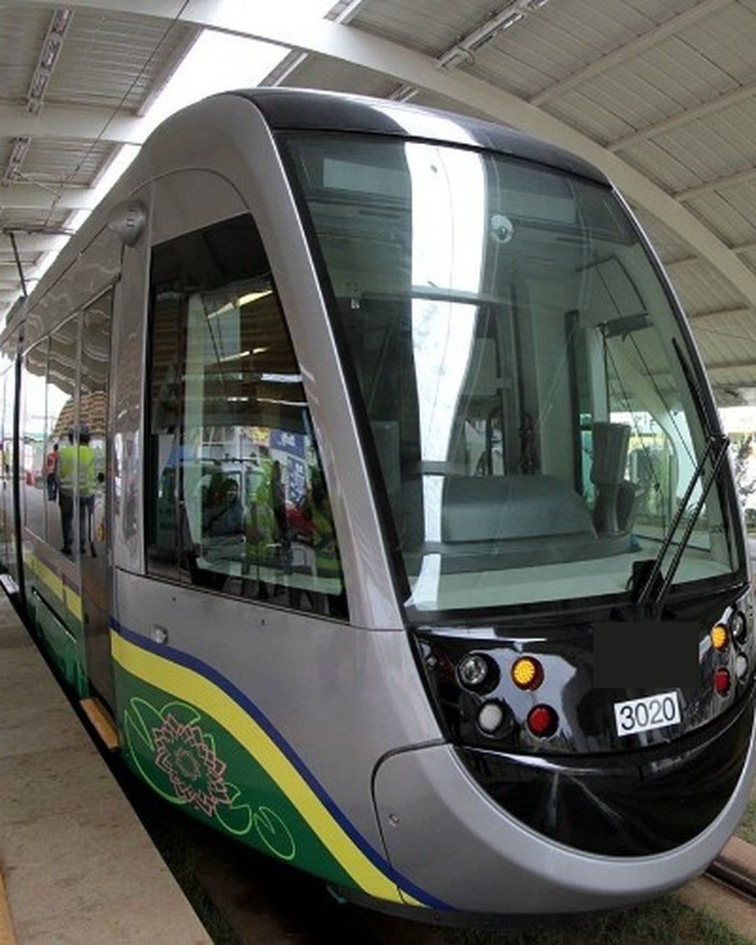 Frente de um trem do VLT prateado, com faixas verde e amarela, parado sob a cobertura de uma estação