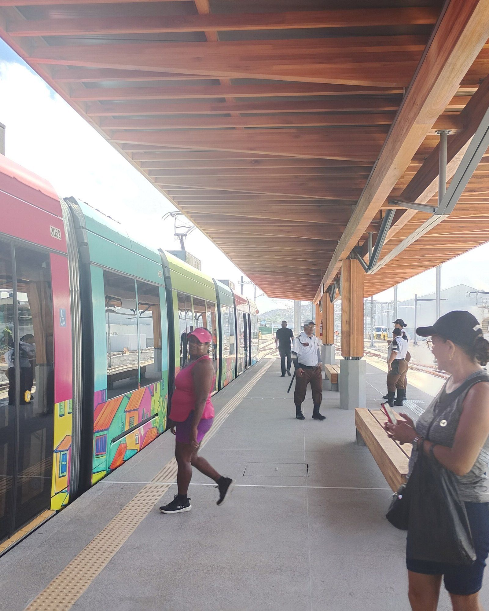 Trem do VLT com pintura colorida de casas parado na plataforma, com passageiros e seguranças sob uma cobertura de madeira